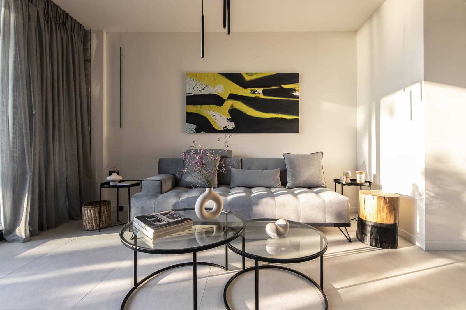 Een moderne zithoek met een grijze bank, twee ronde glazen tafels, een groot abstract schilderij en een houten bijzettafeltje.