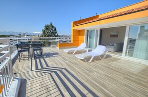 Una terraza con tumbonas y una mesa, una puerta de vidrio que da a un espacio interior, y una vista despejada de montañas al fondo.