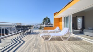 Eine Terrasse mit Liegestühlen und einem Tisch, mit Blick auf ein Gewässer und Gebäude.