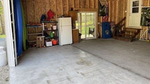 Eine Garage mit Überdachung, Betonboden, Holzwänden, einem Kühlschrank, Regalen mit verschiedenen Gegenständen und einer Flagge.