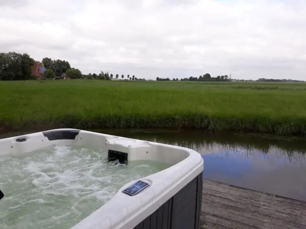 A hot tub with a control panel, surrounded by a wooden deck and a calm water body, with a grassy field and trees in the background.