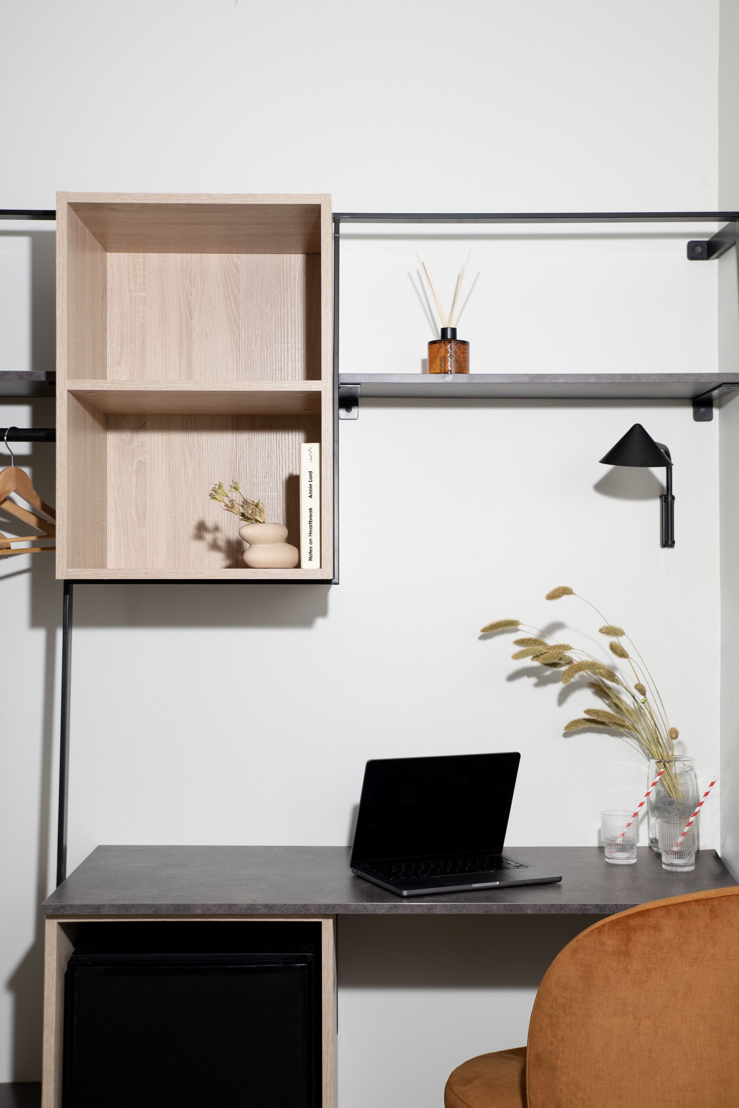 Un espacio de trabajo moderno con una computadora portátil sobre un escritorio, una estantería de madera con objetos decorativos y una lámpara negra.