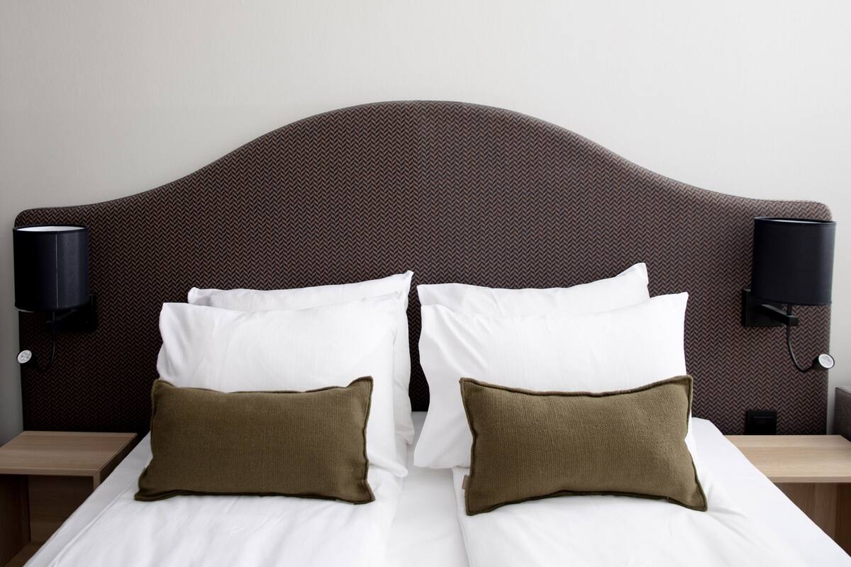 Una cama bien hecha con almohadas blancas y marrones, un cabecero oscuro y texturizado, y dos lámparas de pared a cada lado.