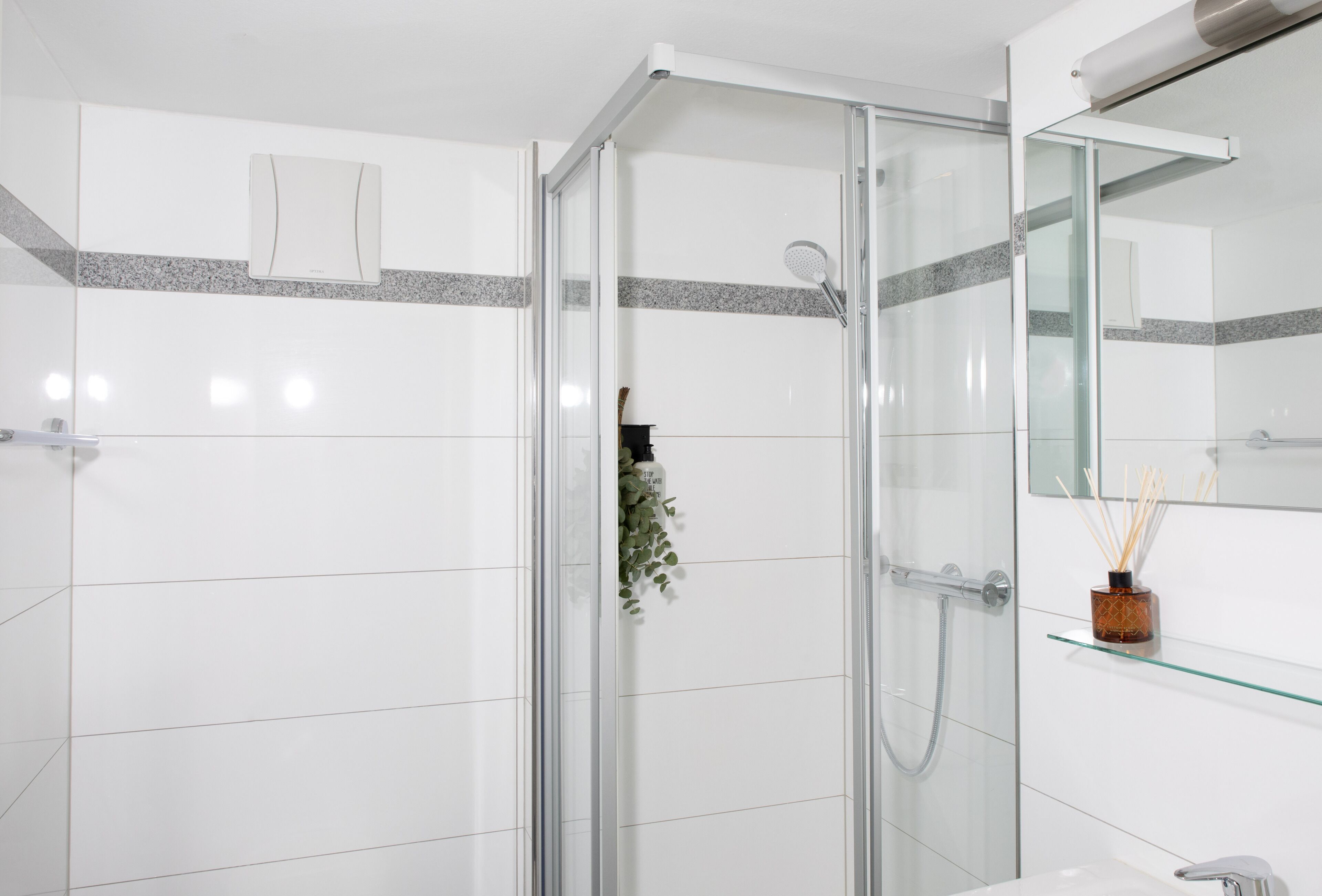 Un baño moderno con azulejos blancos, cabina de ducha de vidrio, repisa de vidrio con un objeto decorativo y un espejo.