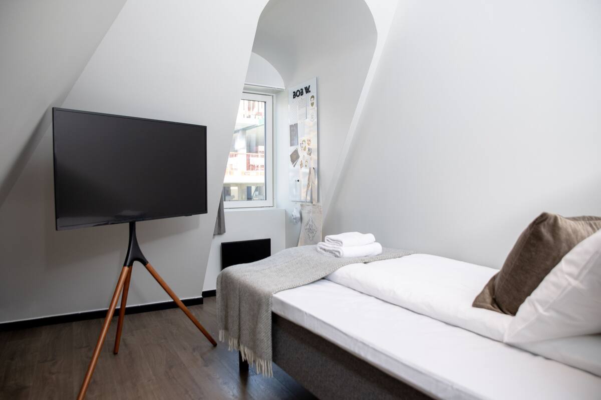 Un dormitorio con cama, un televisor de pantalla plana sobre un atril, una ventana con un cartel y vista a la ciudad.