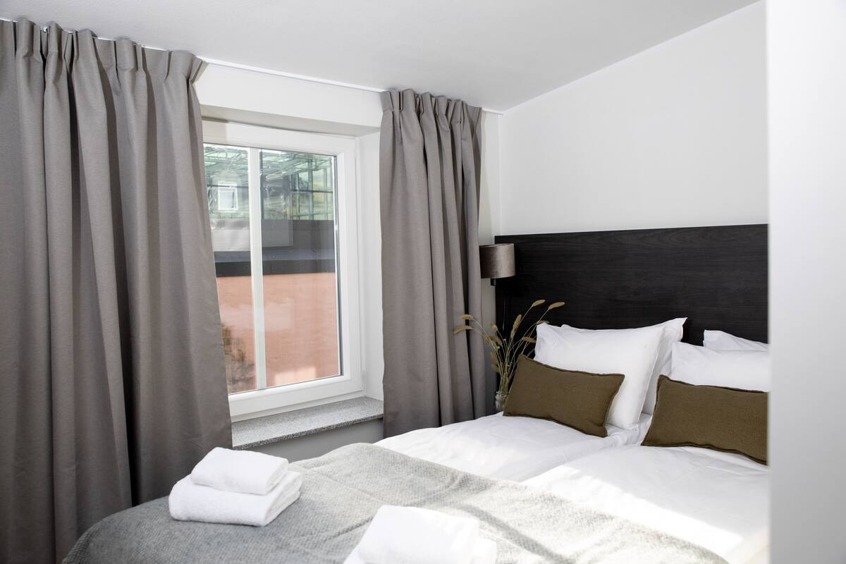 Una habitación de hotel con cama, cortinas, una ventana y una mesita de noche con lámpara.