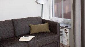 Een donkergrijze bank met een enkel groene kussen en een boek erop, geplaatst naast een vloerlamp en een raam met uitzicht.