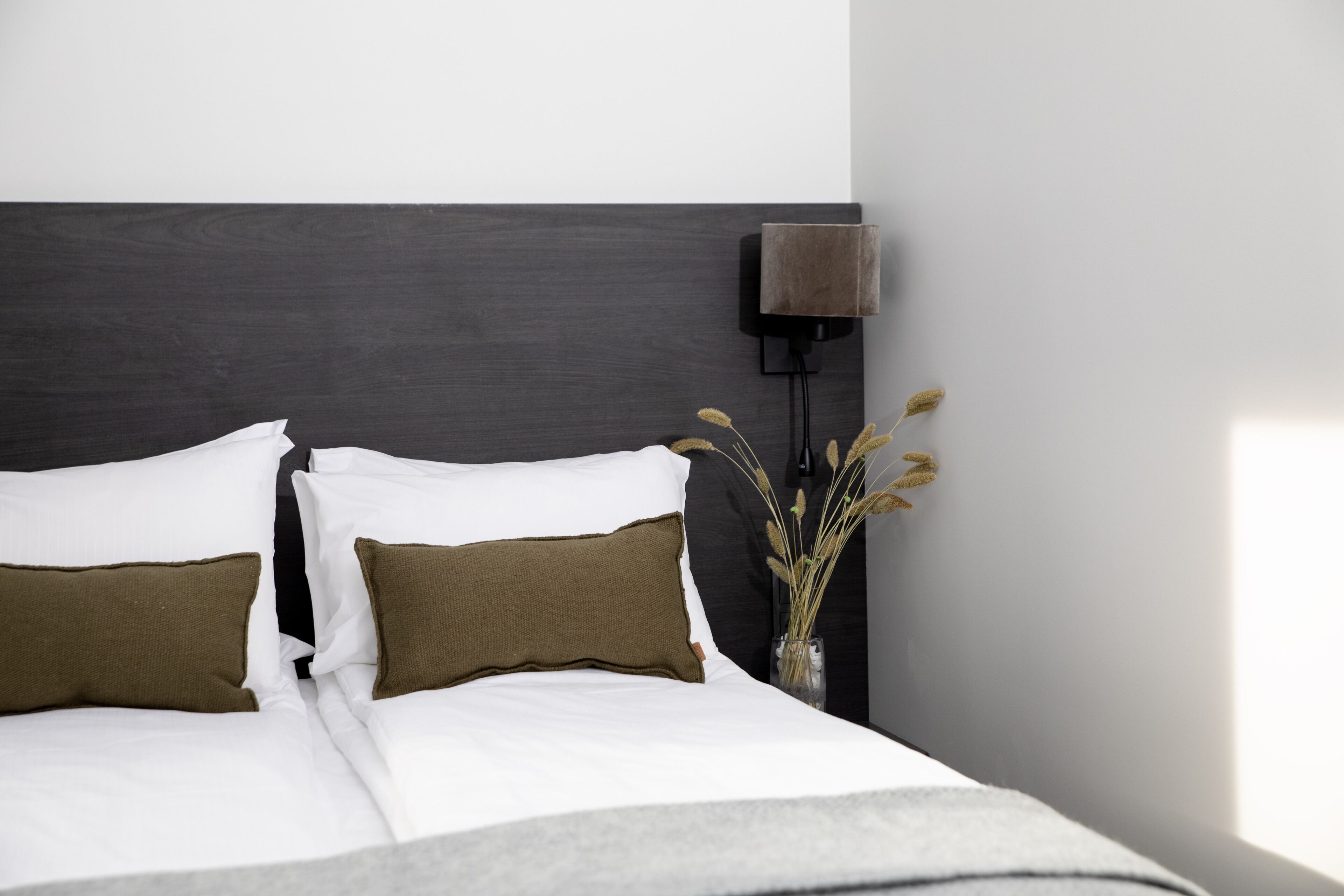 Een netjes opgemaakt bed met wit- en bruin beddengoed, een donker hoofdbord en een bedlampje met gedroogde planten.