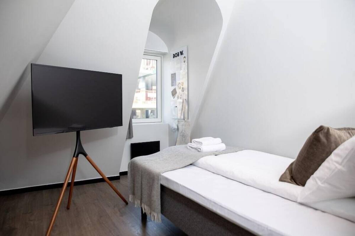 Un dormitorio con una cama, un televisor sobre un atril, una ventana y una pared con un cartel.