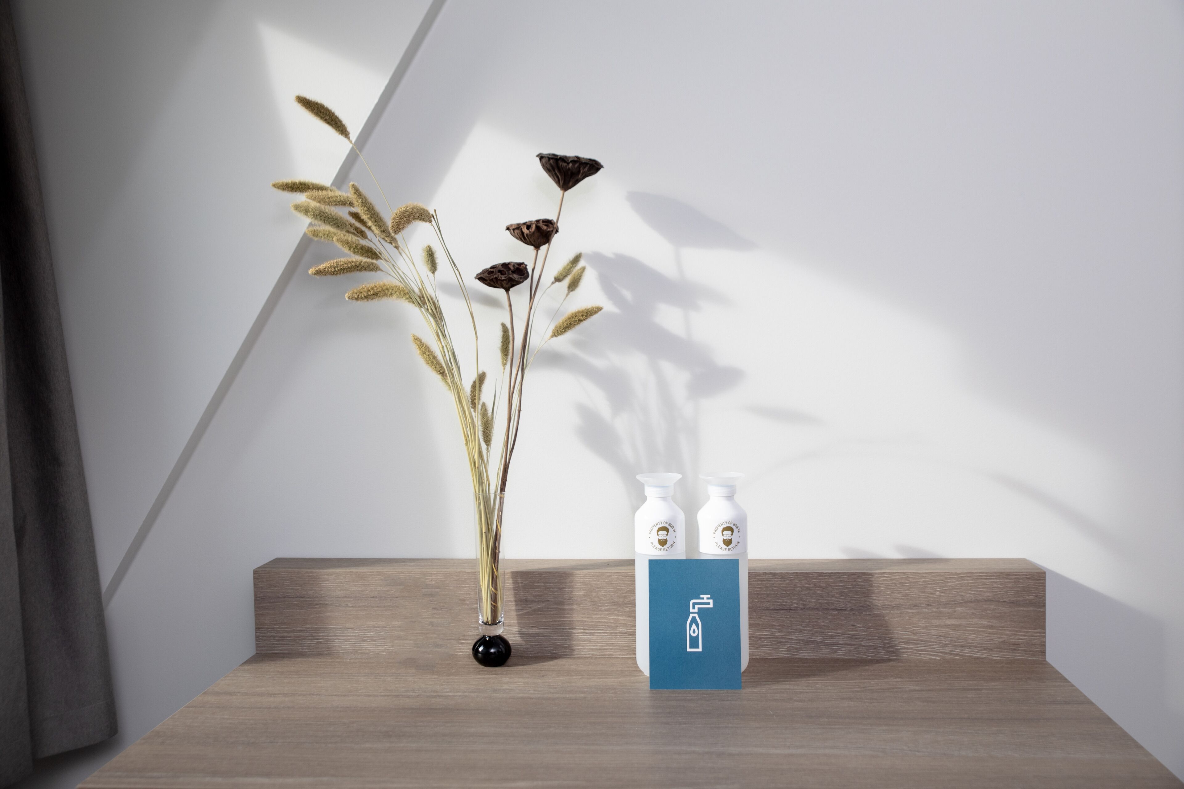 Un estante de madera con un jarrón de plantas secas y dos botellas blancas con etiqueta azul.