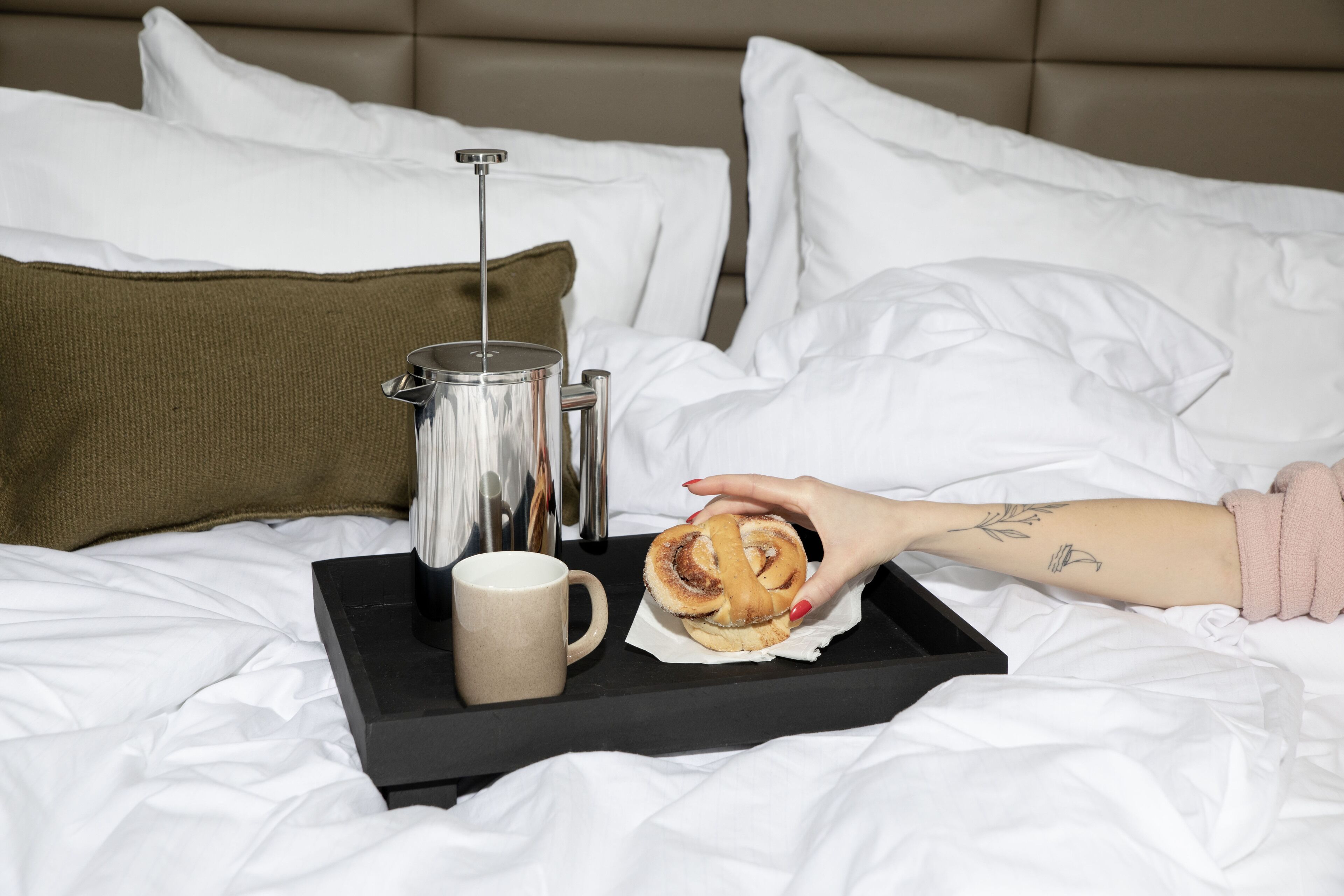 Una cama con sábanas blancas, una bandeja negra con una taza, un pastel y un termo.