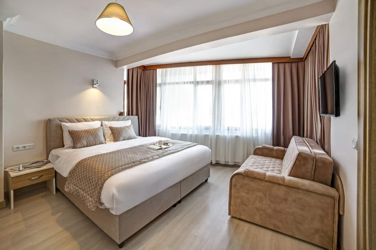 Quarto de hotel moderno com uma cama grande, um sofá, uma televisão de tela plana e uma janela com cortinas.