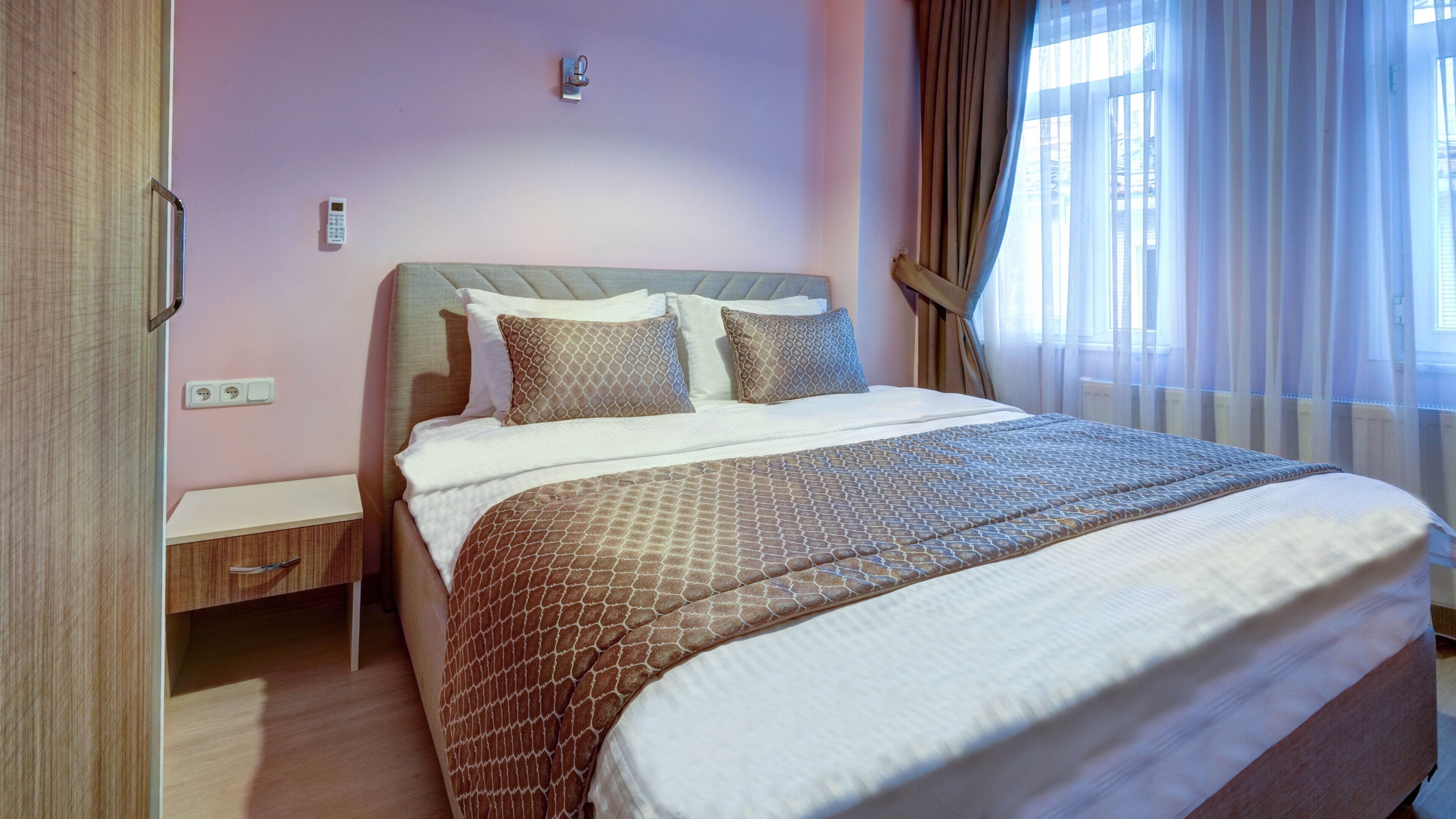 A neatly made bed with patterned bedding, a bedside table, a wall-mounted light, and a window with curtains.