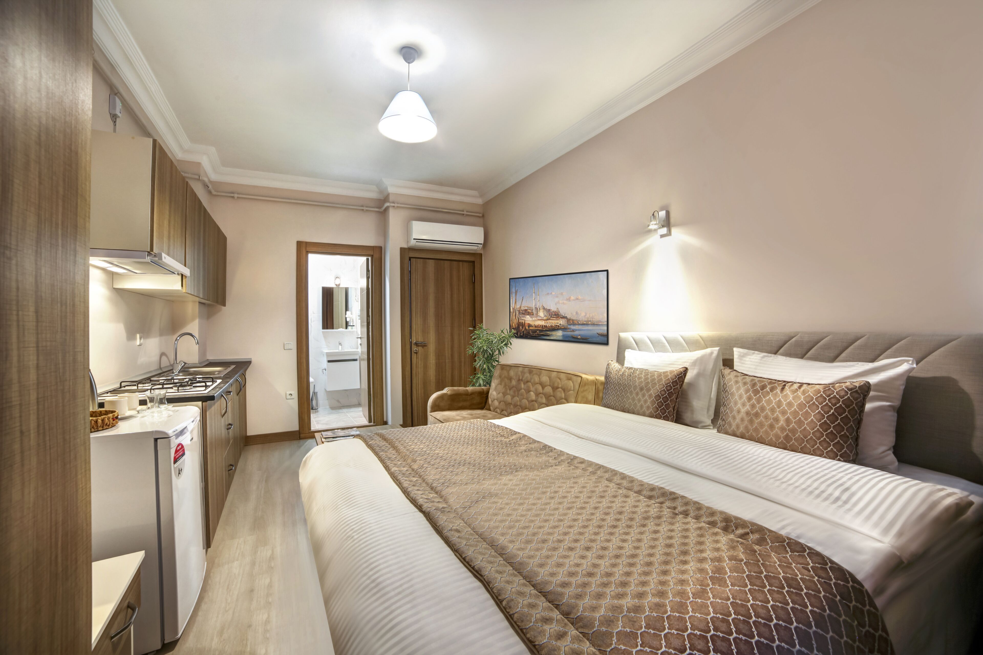 A hotel room with a bed, a desk, a chair, a small kitchenette, and a door leading to a bathroom.