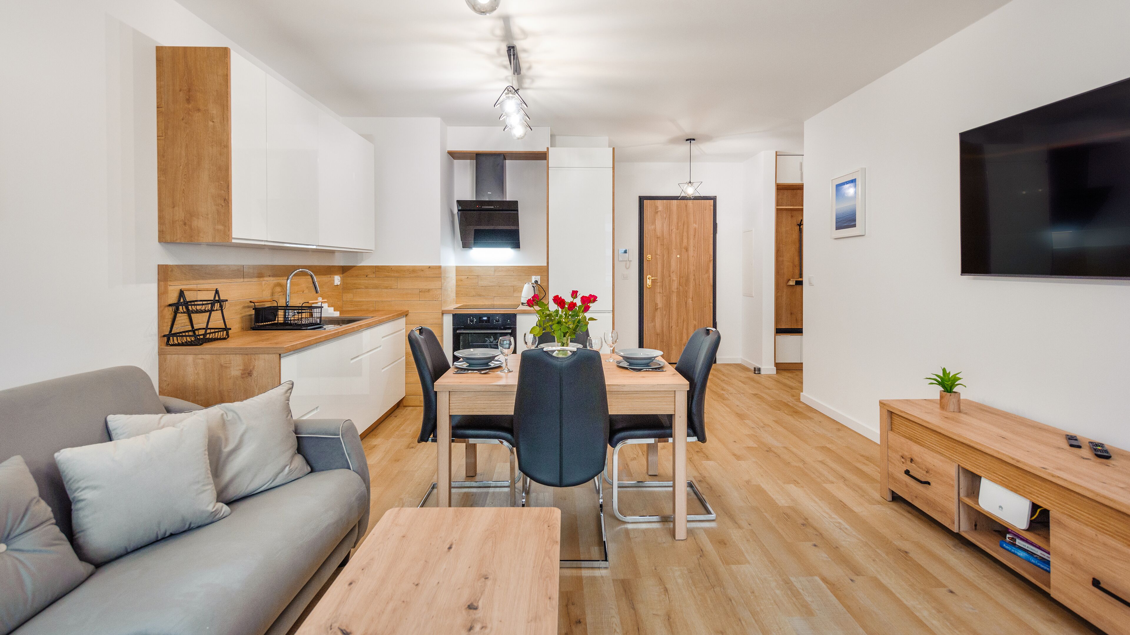 A modern, well-lit living space with a dining area, kitchen, and a flat-screen TV mounted on the wall.