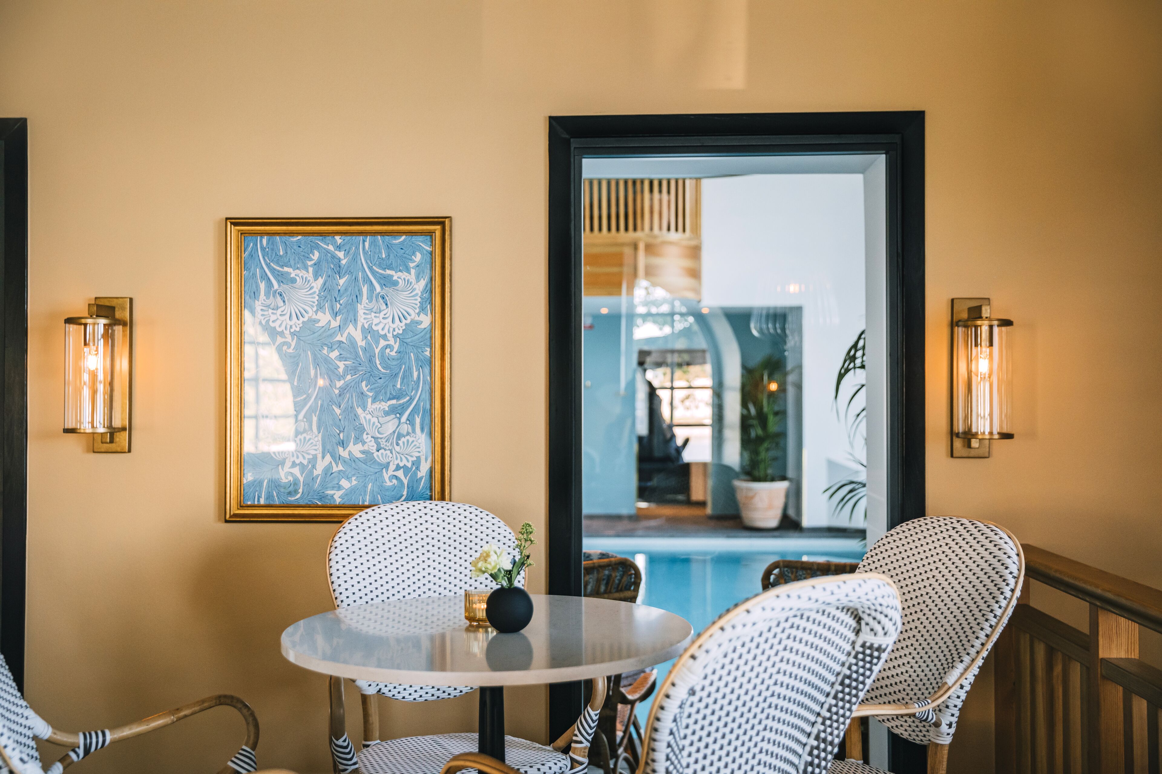 Un coin repas avec une table ronde et des chaises en osier, une œuvre d’art encadrée au mur et, vue à travers une porte, une piscine.
