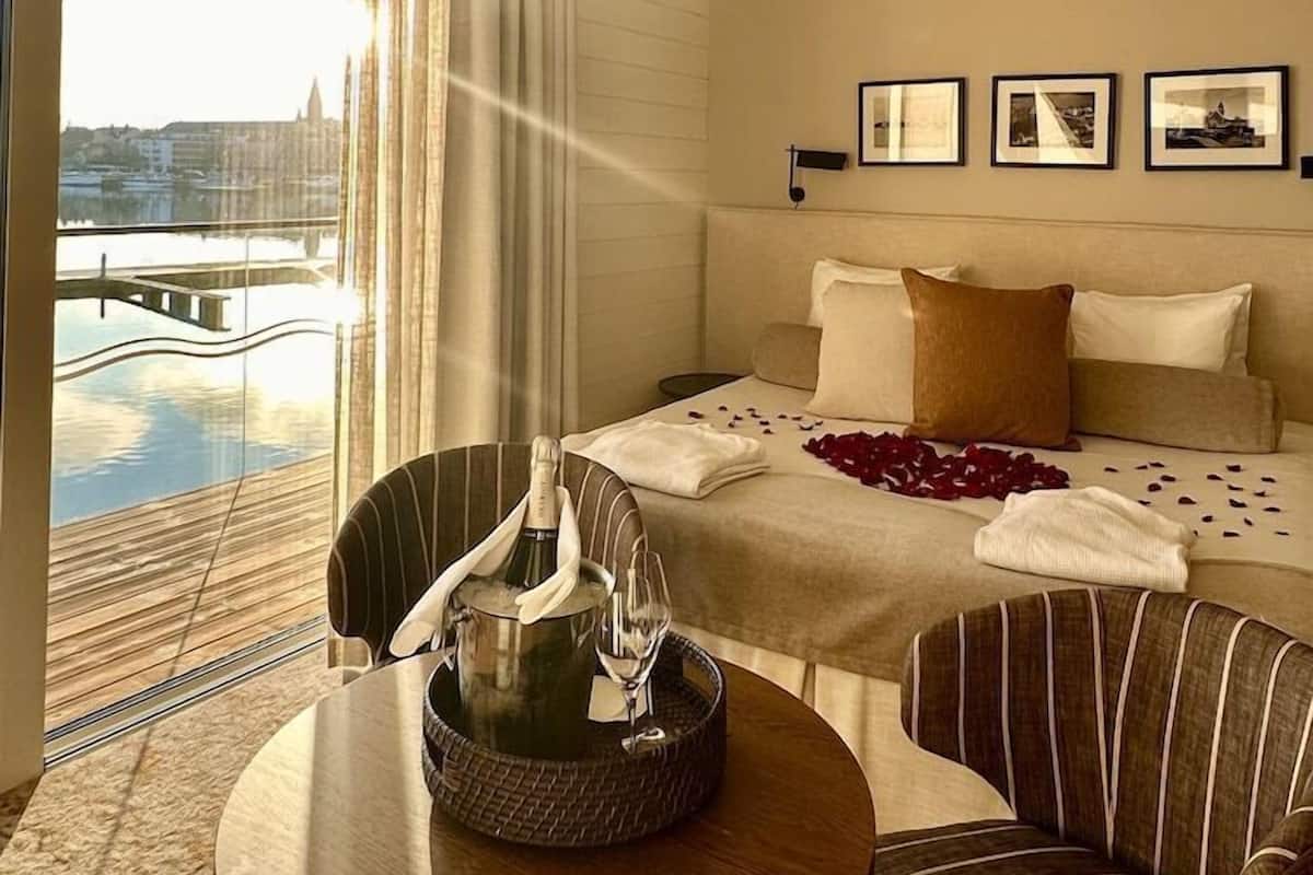 A hotel room with a bed, a dining table with a bottle and glasses, a chair, and a view of a body of water and buildings.