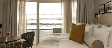 Habitación de hotel con cama, una silla, una mesa y vistas a una masa de agua a través de amplias ventanas.