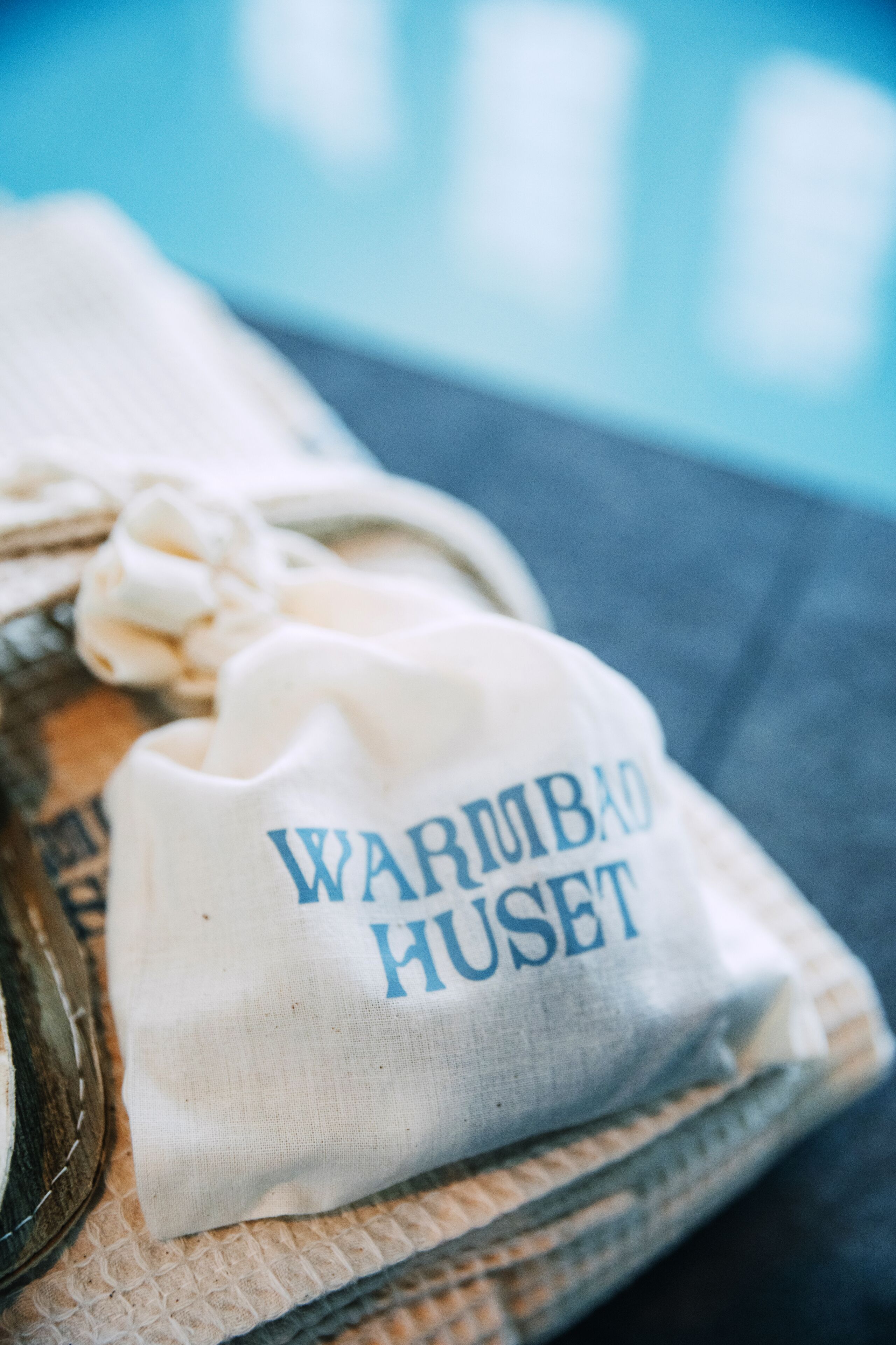 A white drawstring bag with "WARM BAD HUSET" printed on it, placed on a textured surface near a pool.