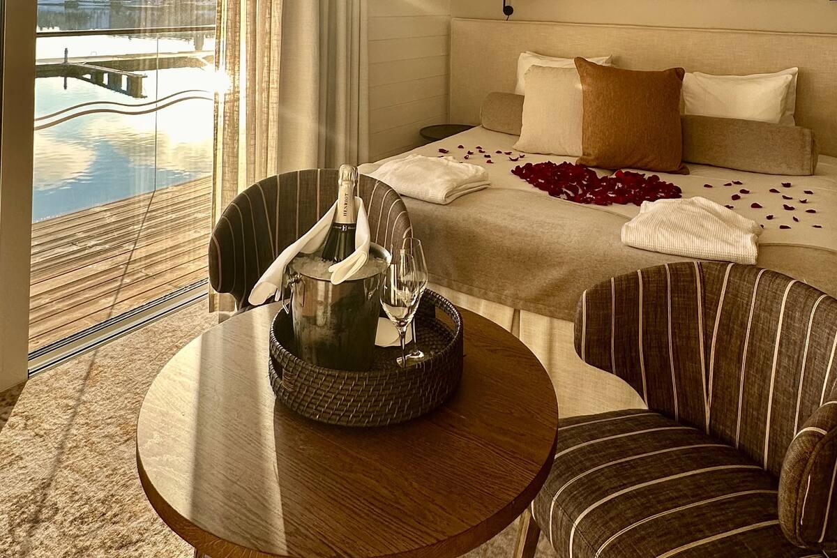A hotel room with a bed, a dining table, a chair, and a view of a cityscape through a large window.
