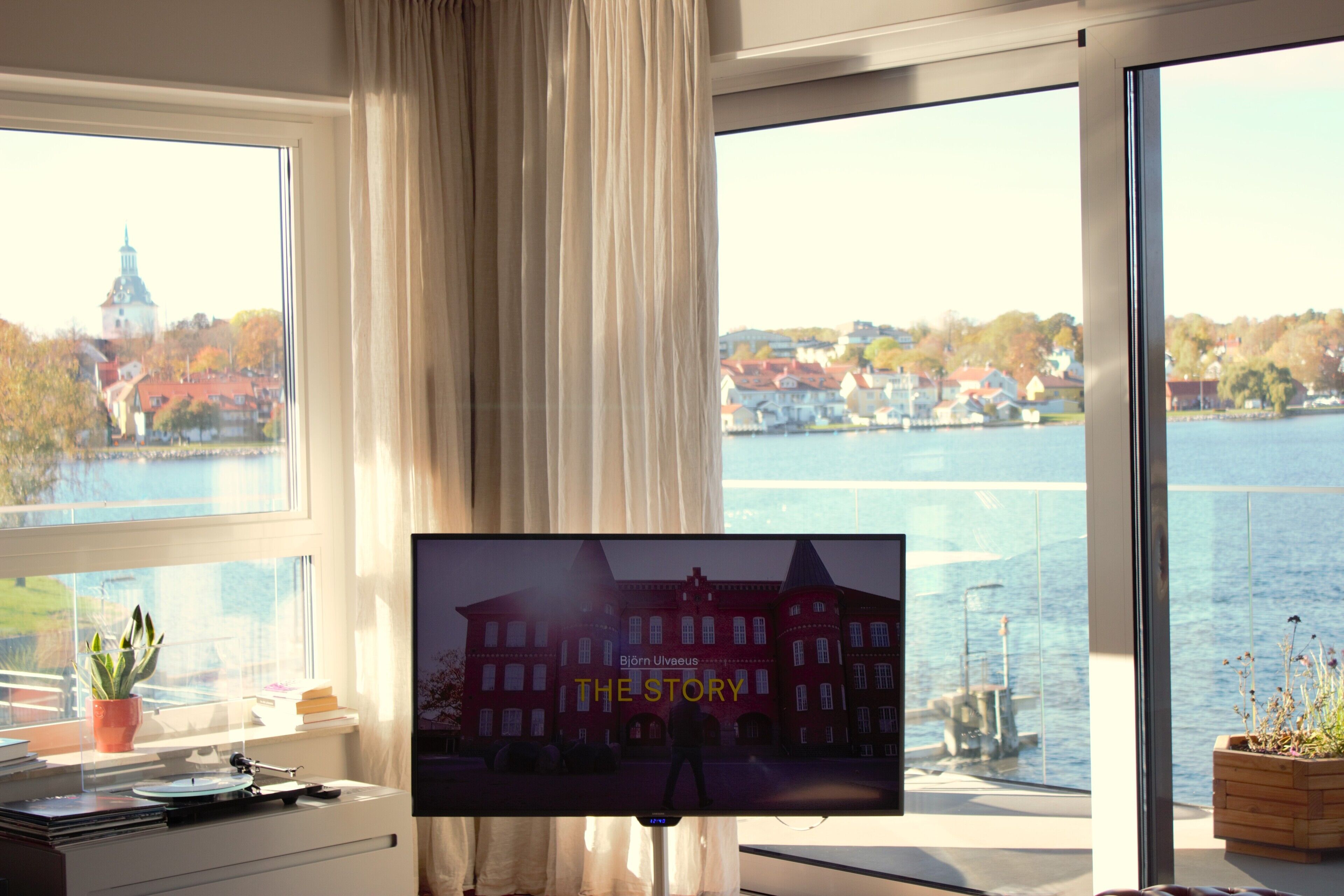 A room with a large window overlooking a body of water and a cityscape, a television displaying 'THE STORY' on the screen, and a plant on the windowsill.