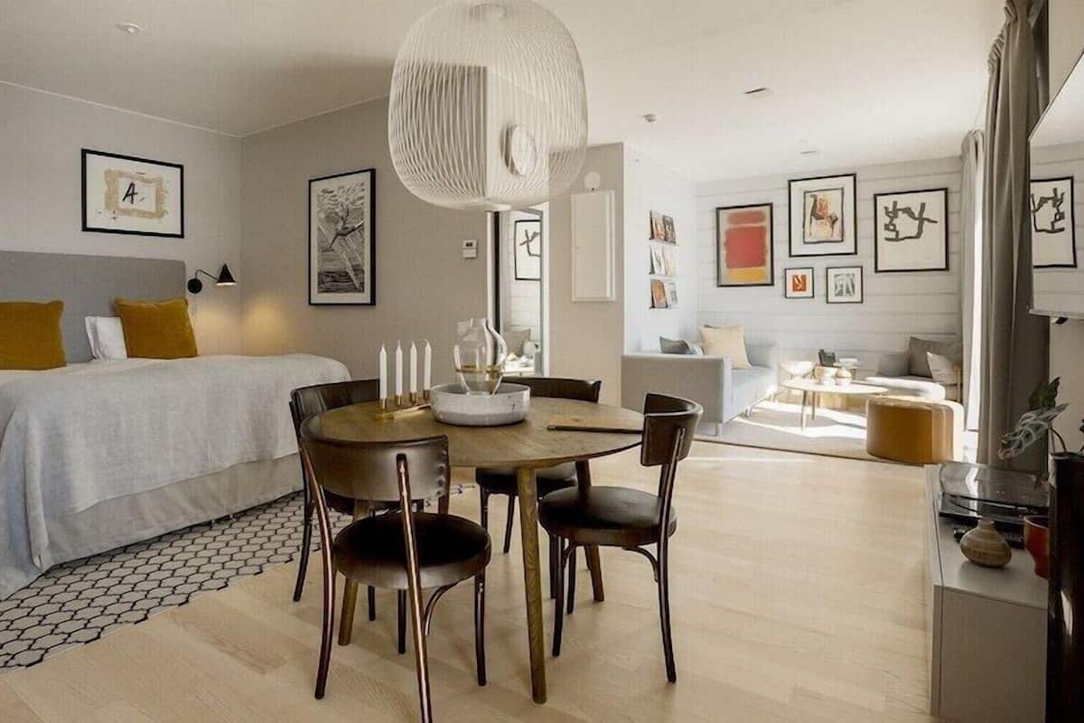 A modern hotel room with a bed, bedside table, lamp, and a dining area with chairs and a table.