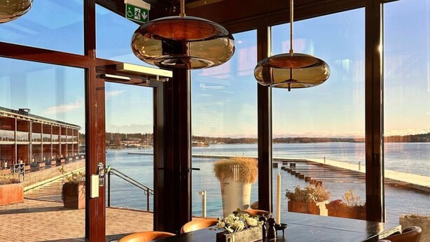 A bar with a view of a waterfront, featuring a long counter, orange chairs, and hanging pendant lights. - Slottsholmen hotell och restaurang (Vastervik)
