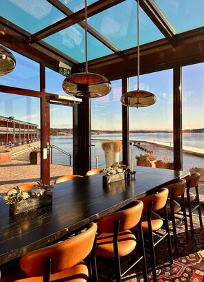 A bar with a view of a waterfront, featuring a long counter, orange chairs, and hanging pendant lights. - Slottsholmen hotell och restaurang (Vastervik)