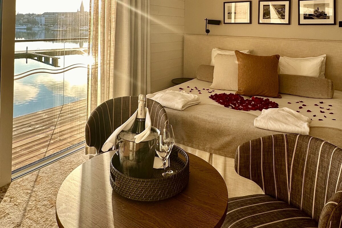 A hotel room with a bed, a table with a bottle and glasses, a chair, a window with curtains, and framed pictures on the wall.