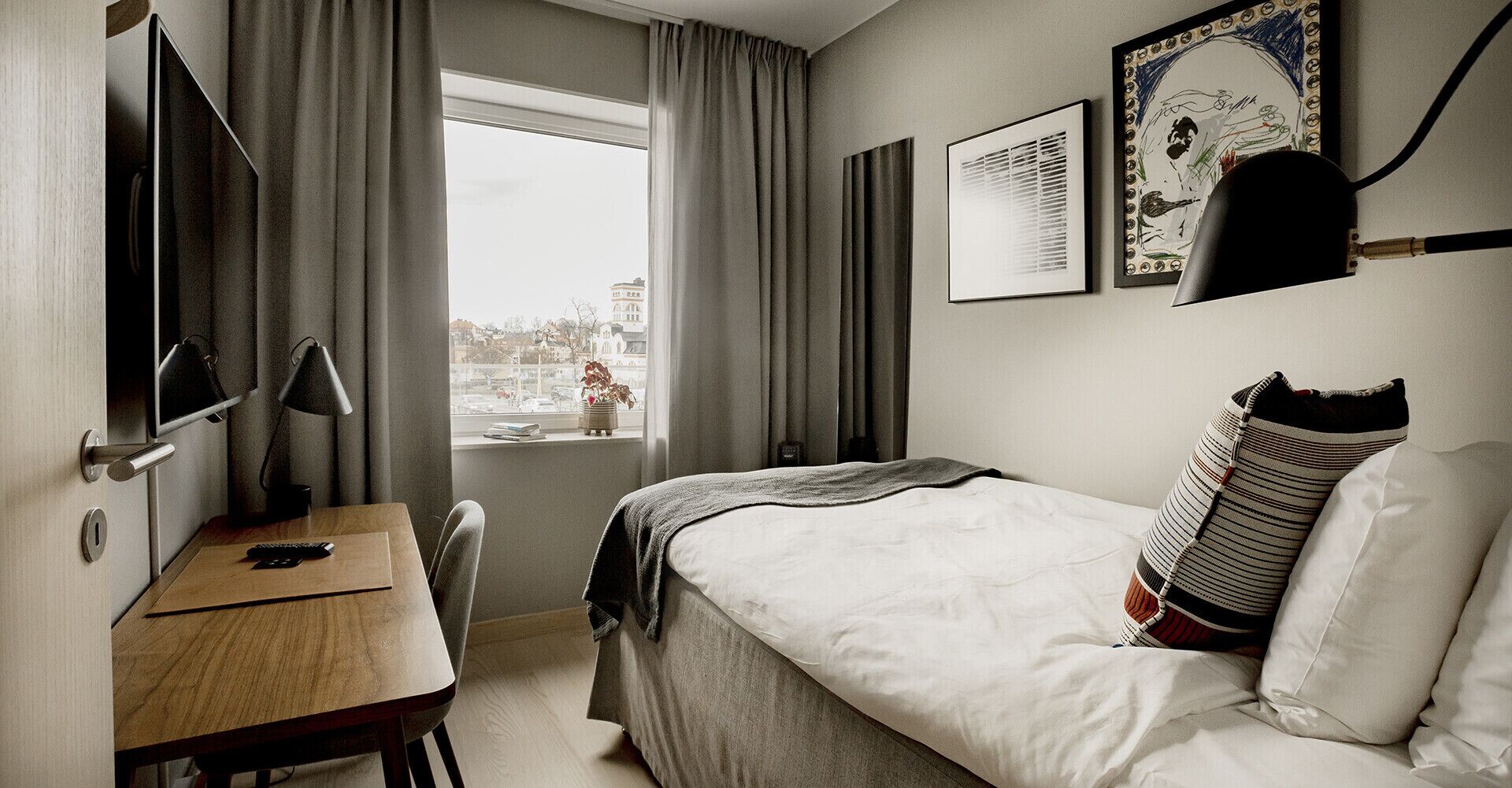 Ett hotellrum med en säng, ett skrivbord, en stol, ett fönster med gardiner och konstverk på väggen.