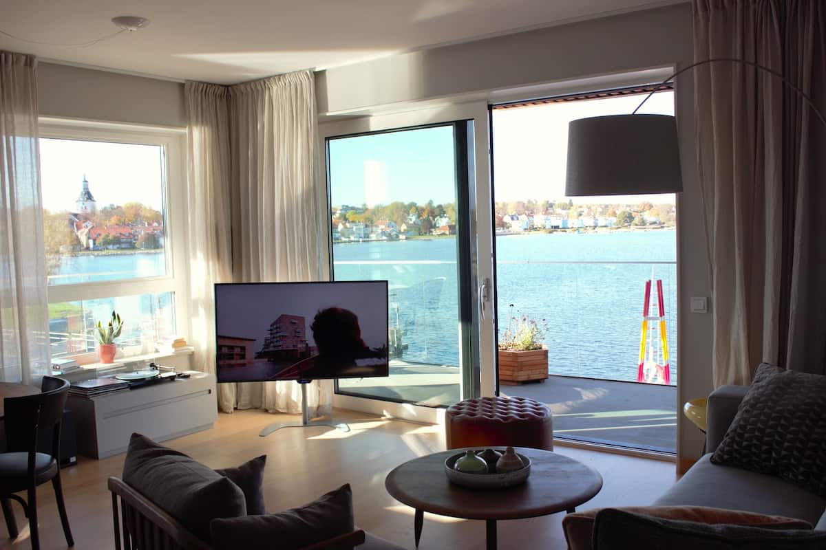A modern living room with a large TV, comfortable seating, a small table, and a view of a body of water through large windows.