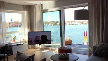 A modern living room with a large TV, comfortable seating, a small table, and a view of a body of water through large windows.