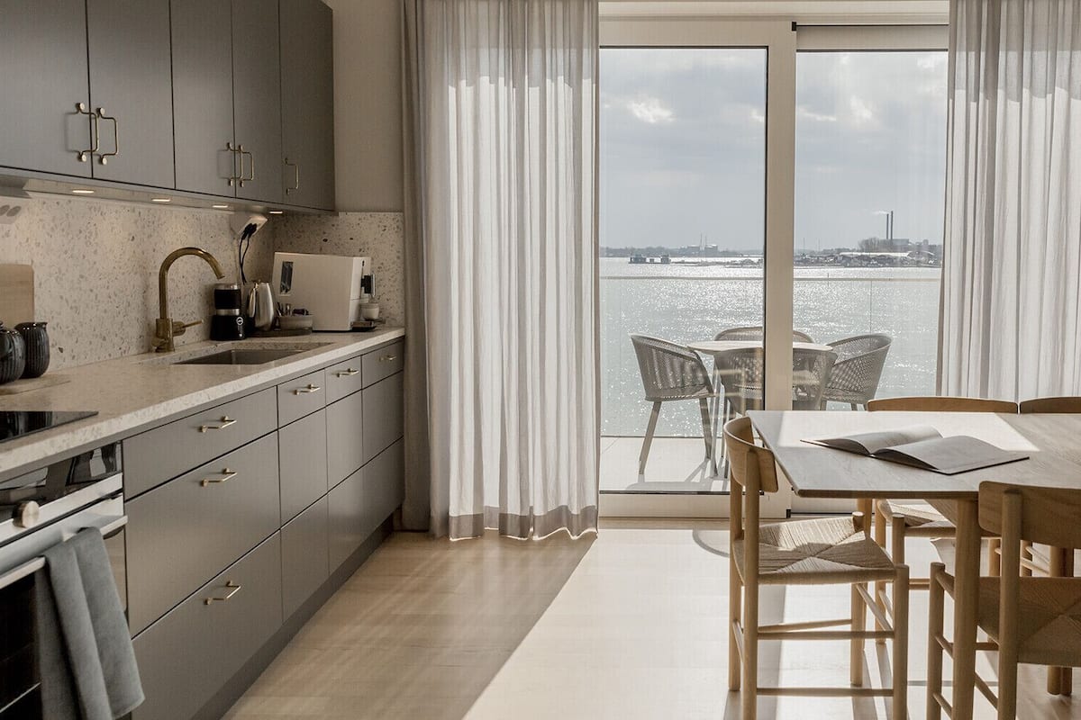 A modern kitchen with a large window overlooking a body of water, a dining area with wooden furniture, and a view of the water.