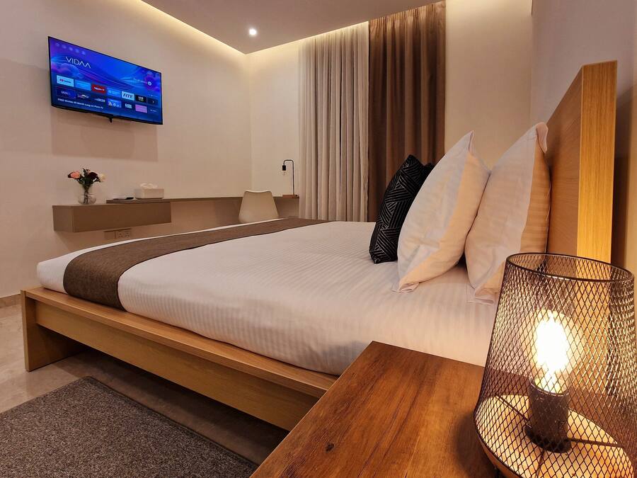 Quarto de hotel moderno com uma cama grande, uma TV fixada na parede, uma mesinha de cabeceira com luminária e um criado-mudo de madeira.