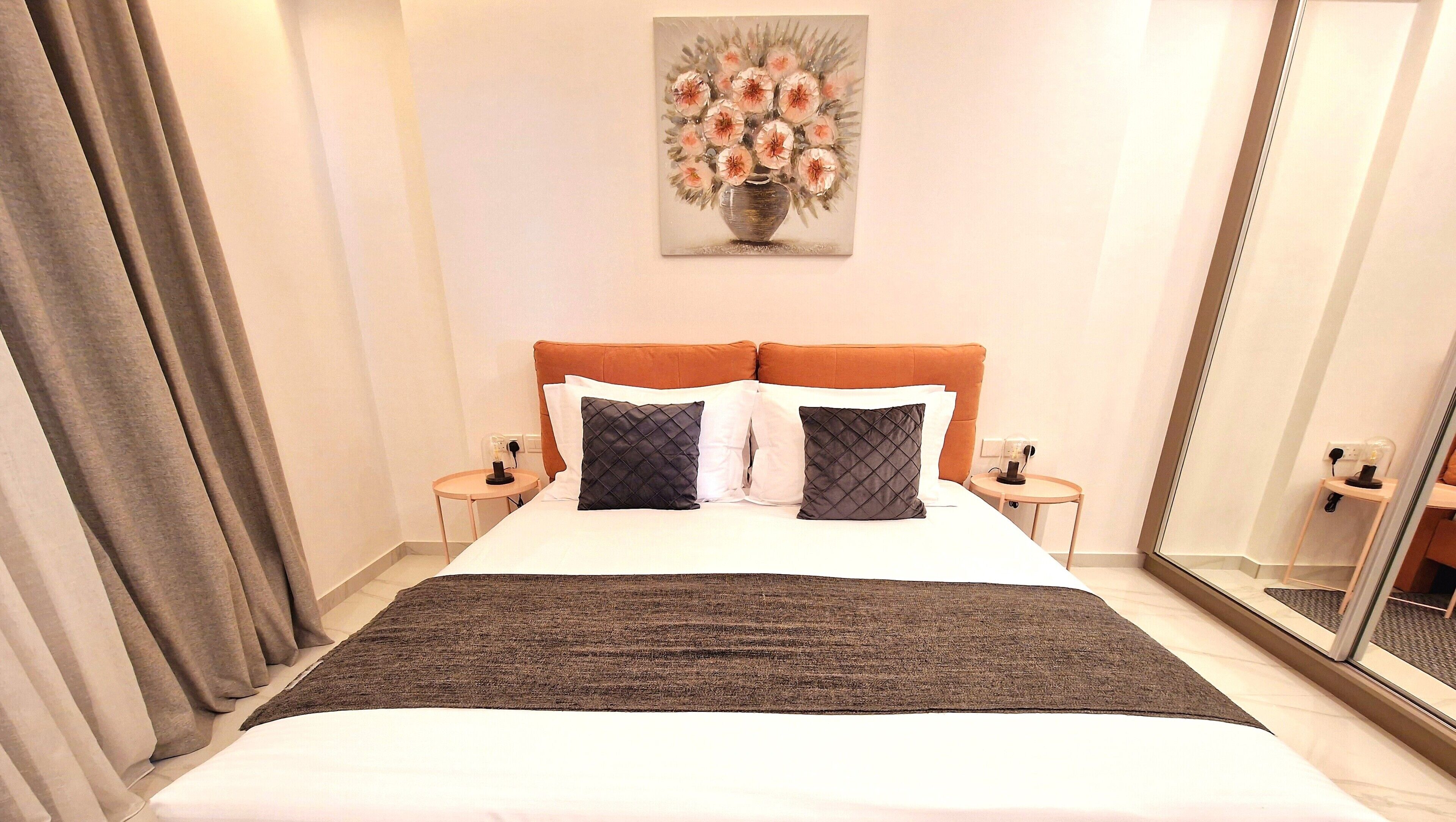 Quarto de hotel com uma cama grande, duas mesinhas de cabeceira, um criado-mudo, um espelho e um quadro na parede.