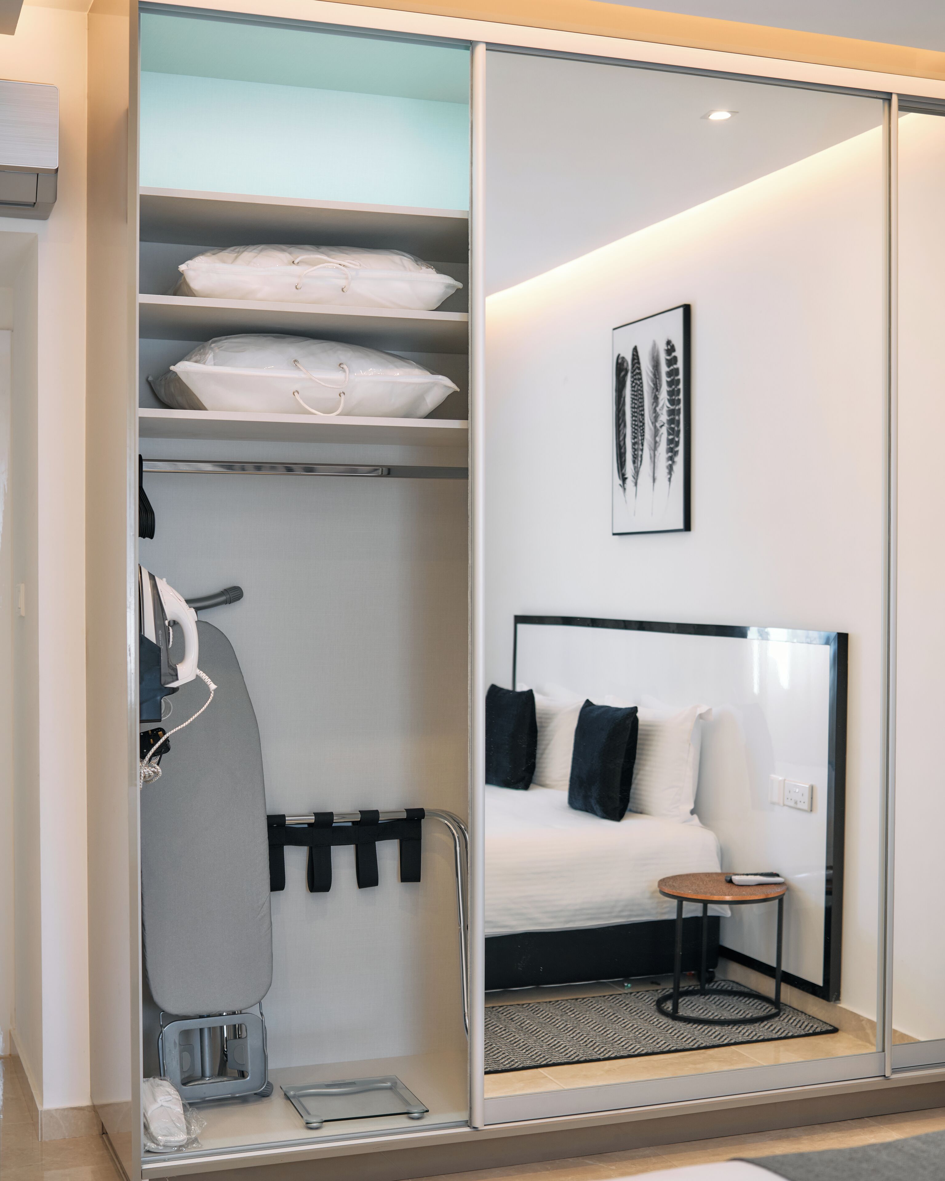 A modern hotel room with a walk-in closet, a bed with black and white bedding, a small round table, and a framed artwork on the wall.
