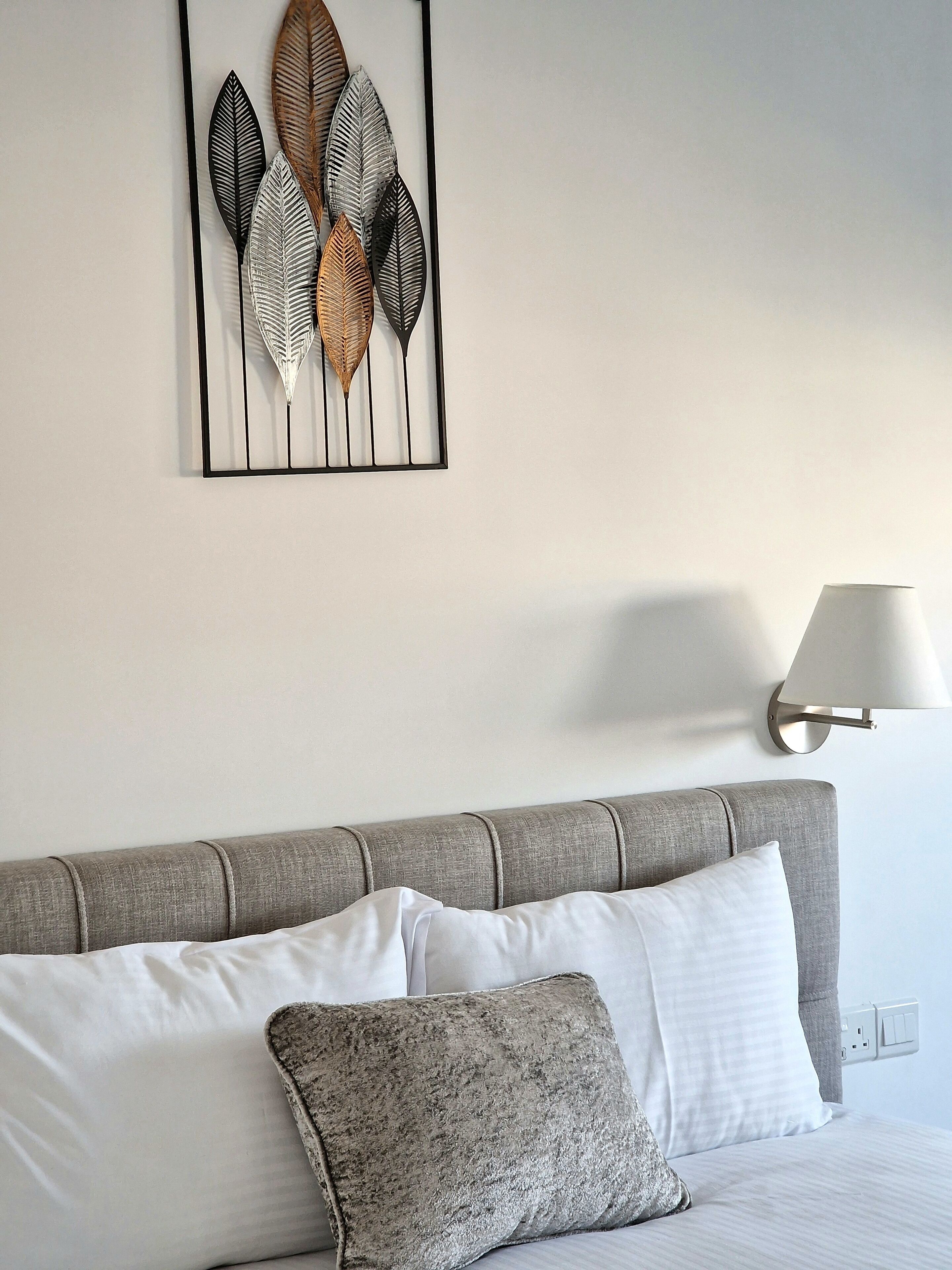 A neatly made bed with white and gray bedding, a gray headboard, and a wall-mounted lamp.