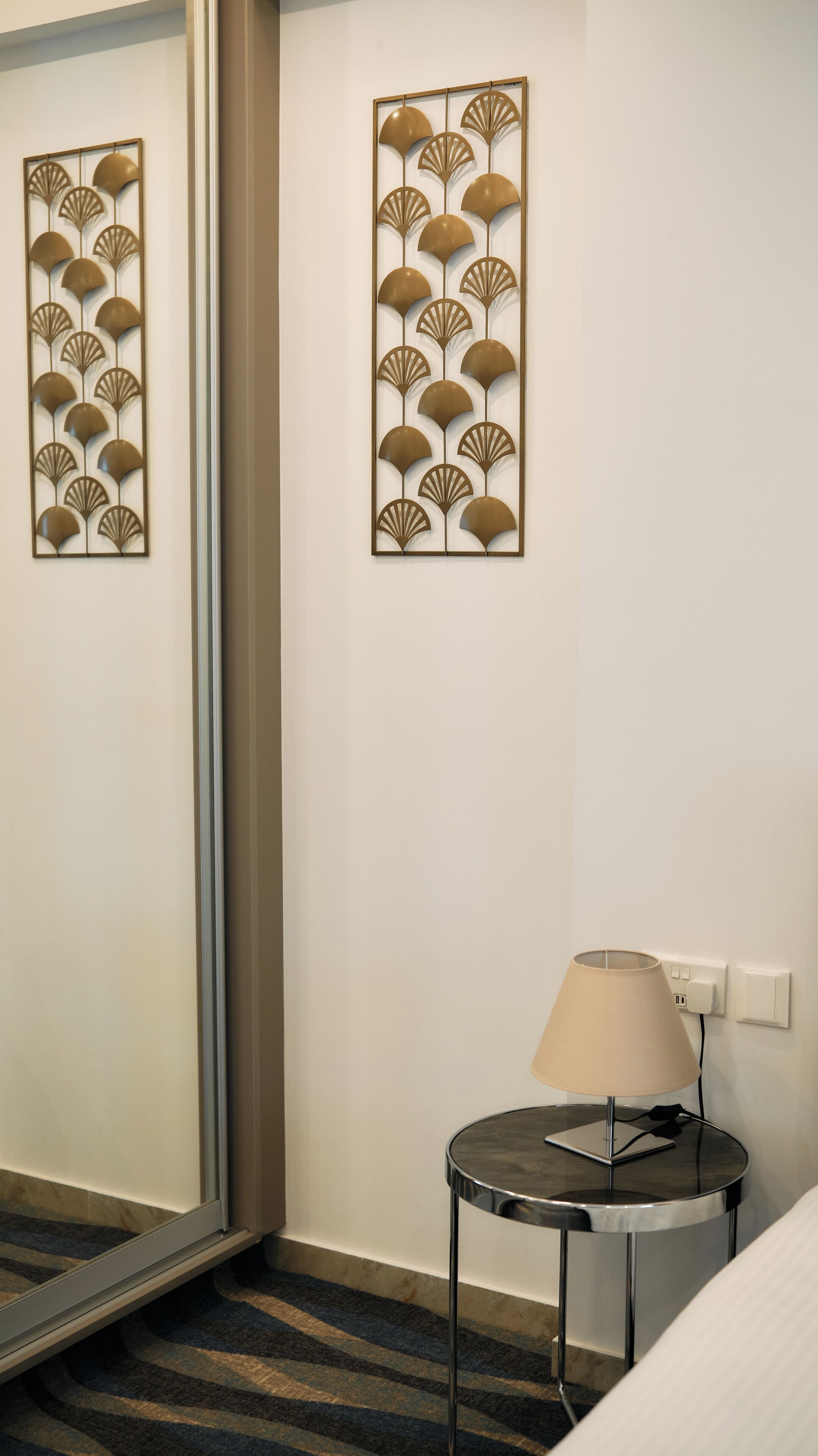 A hotel room with a bed, a round table with a lamp, and two wall-mounted decorative panels.