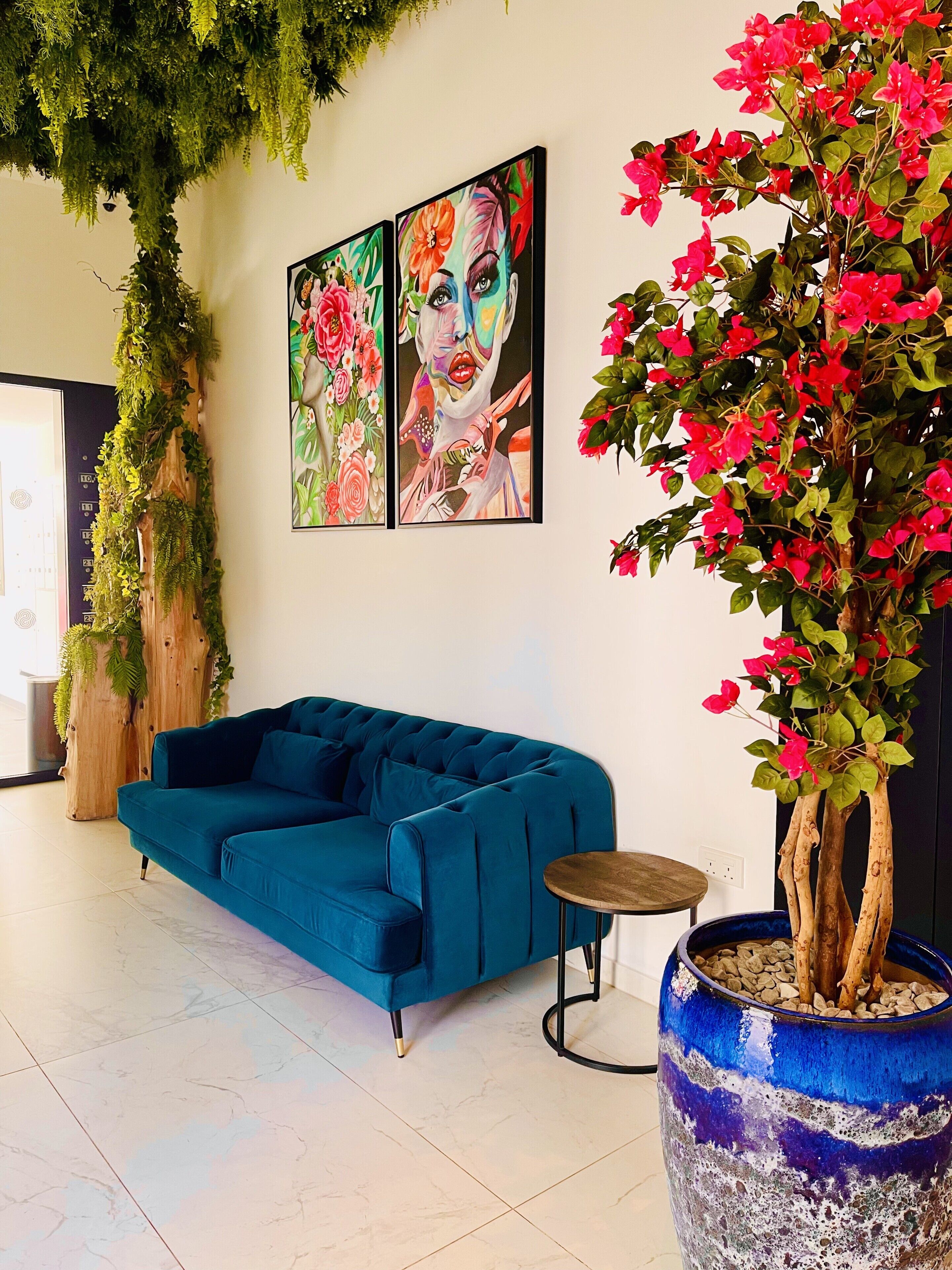 Un soggiorno moderno con un divano color petrolio, un tavolo rotondo in legno, un vaso azzurro di grandi dimensioni con un albero e due opere d'arte incorniciate appese al muro.
