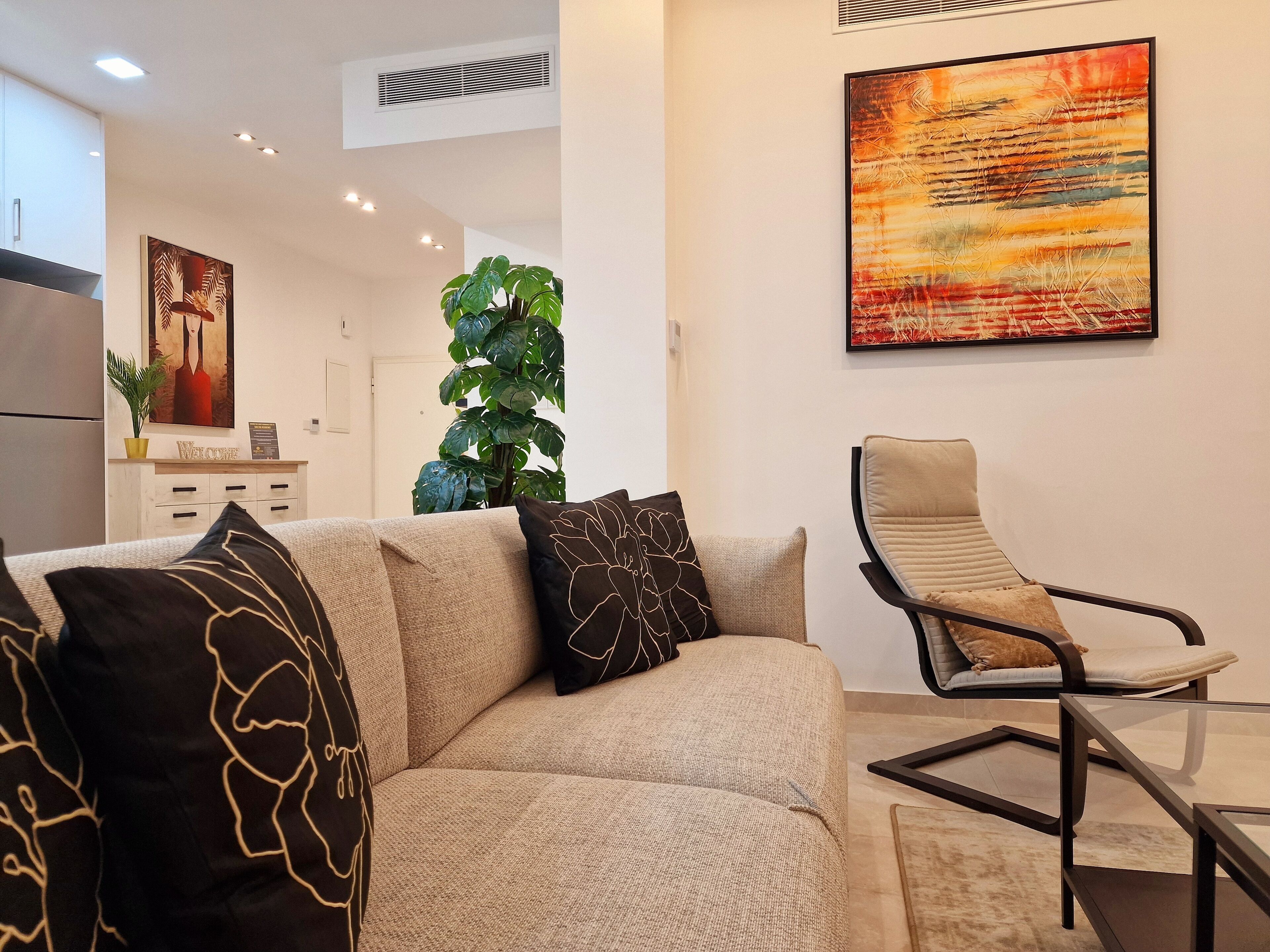 Een moderne woonkamer met een beige bank, een glazen salontafel en een zwarte metalen stoel. Aan de muur hangt een groot abstract schilderij en in de hoek staat een potplant.