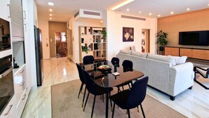A modern living room with a sofa, TV, and dining area with chairs and a table. - Sveltos Residence 3Bedroom Suites (Larnaca)