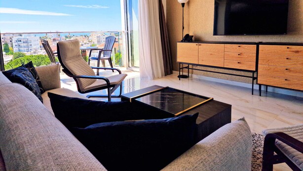 A modern living room with a sofa, armchair, and coffee table, a TV mounted on the wall, and a balcony with a view of the city. - Sveltos Residence 3Bedroom Suites (Larnaca)