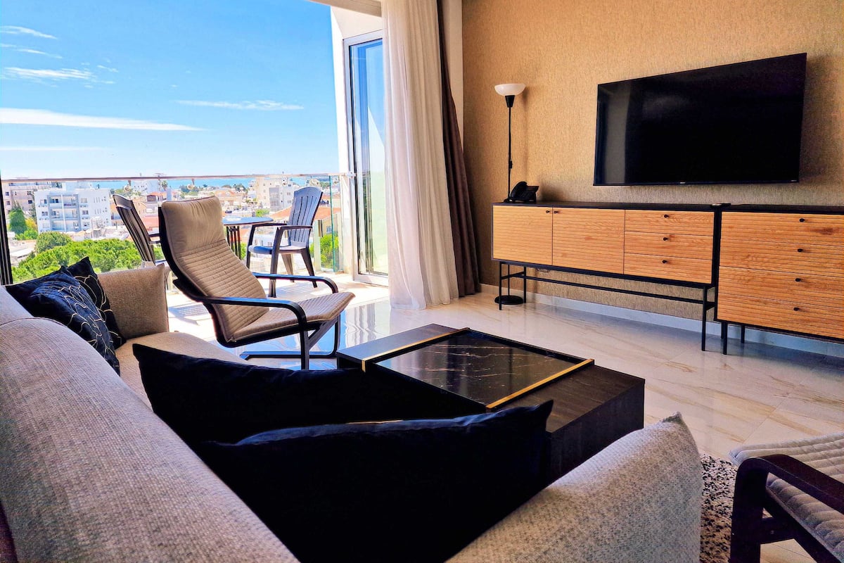 A modern living room with a sofa, armchair, and coffee table, a TV mounted on the wall, and a balcony with a view of the city.