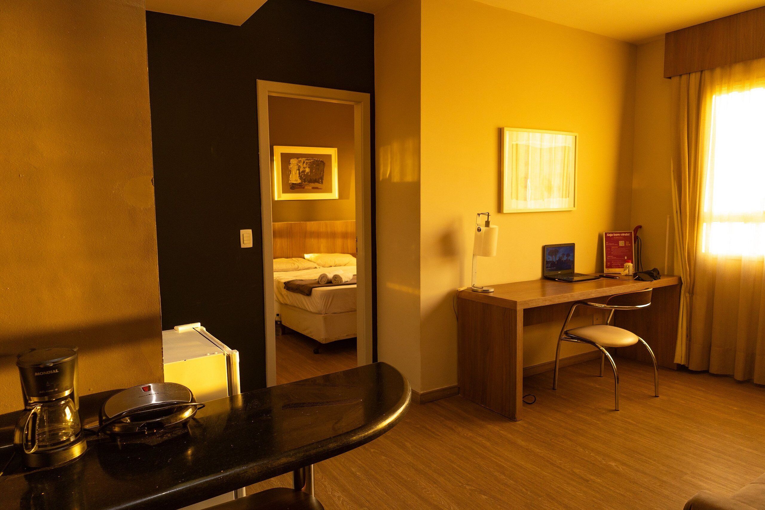 Quarto de hotel com cama, secretária, cadeira, cafeteira e computador.