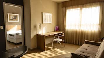 A modern hotel room with a wooden desk, a comfortable sofa, a bed with white linens, and a large window with sheer curtains.