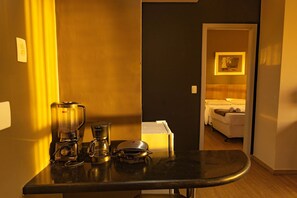 A kitchen counter with a blender, coffee maker, and a small bowl. A door leads to a bedroom with a bed and a painting on the wall. - Locking´s Barro Preto 4 (Belo Horizonte)