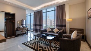 A modern hotel room with a sofa, armchair, and a desk with a chair. There is a large window with curtains, a floor lamp, and a patterned rug. - Gladiolus Hotel (Jeddah)