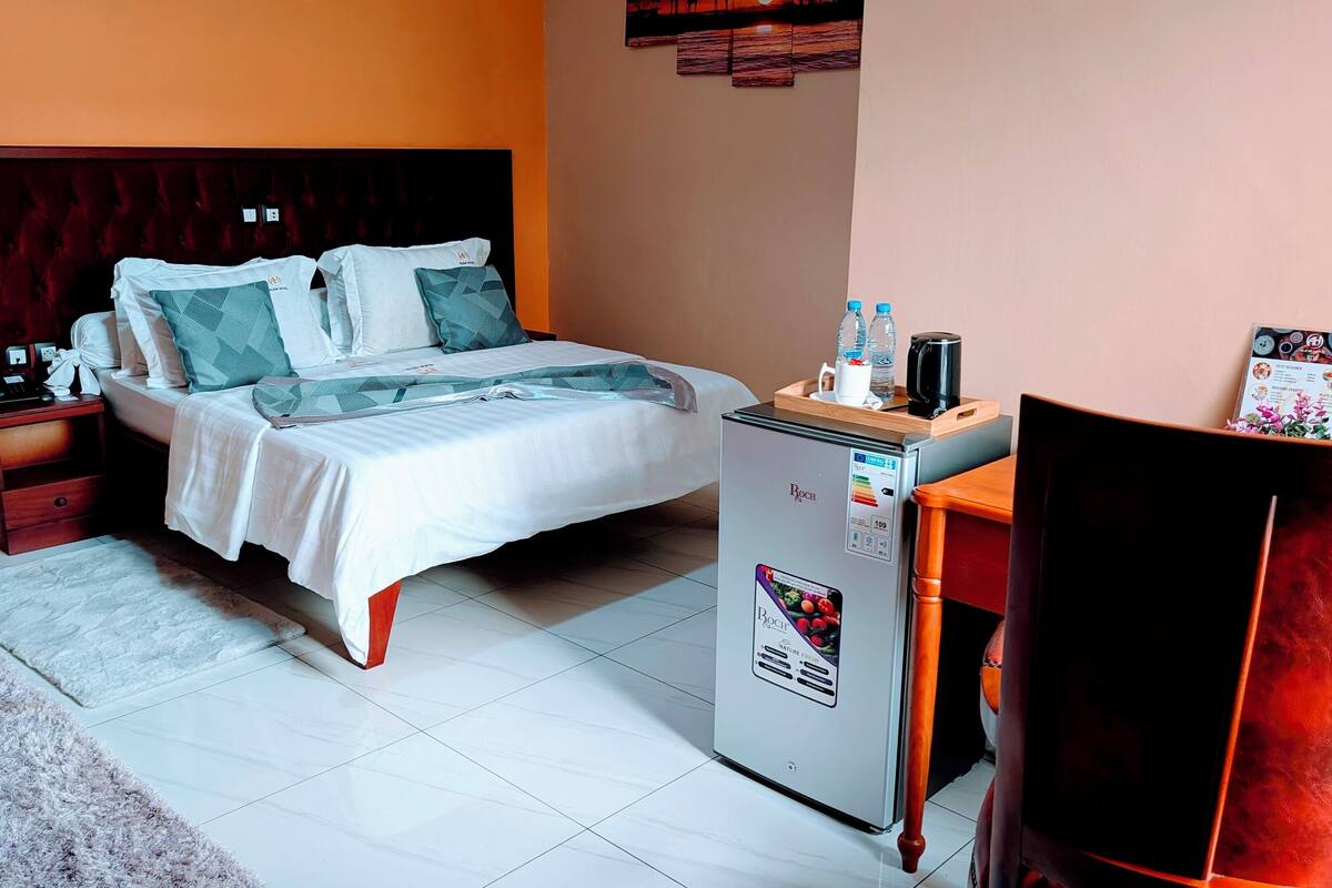 A hotel room with a bed, a small refrigerator, a wooden nightstand, a red chair, and a wall-mounted artwork.