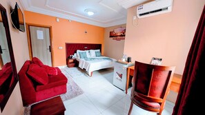 A hotel room with a bed, a desk, a chair, a red sofa, and an air conditioning unit. - Alicia Hotel (Douala)