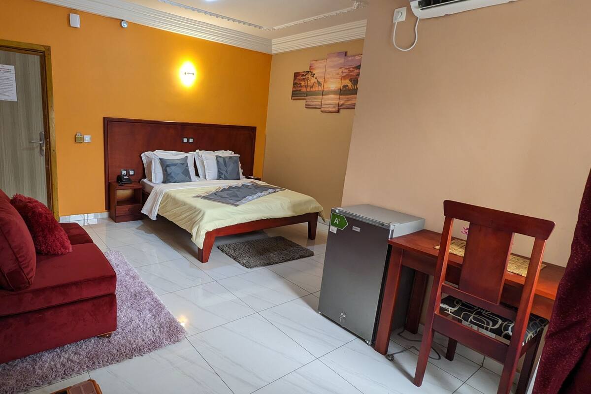 A hotel room with a bed, a red sofa, a small table, and a refrigerator.